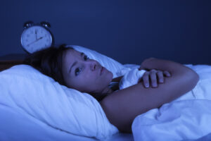 Mujer acostada en la cama con los ojos abiertos durante la noche, mirando al techo con expresión de insomnio, mientras un reloj marca las 3 a. m., representando una escena típica al hablar de cuáles son los trastornos del sueño más comunes, como el insomnio o la dificultad para conciliar el sueño.
